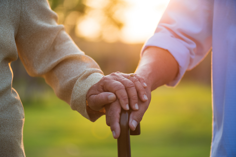 A senior couple holds hands while one uses a cane to walk